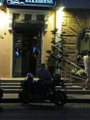 A person is sitting on a motorcycle parked on the street in front of a lit-up bakehouse entrance. The scene takes place at night, with the warm glow of the street lights illuminating the textured stone walls of the building. The entrance features a window displaying reflections, and there are some plants visible near the steps.