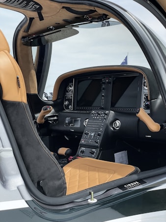 The image captures the interior of a modern aircraft cockpit, featuring sleek and sophisticated controls and displays. The seats are upholstered in tan and black leather with a quilted pattern, and multiple screens and buttons are visible on the console. The setup appears to be designed for a luxurious and high-tech flying experience.