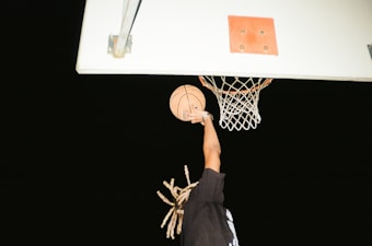 A person is mid-air, performing a slam dunk with a basketball, reaching towards the hoop. The setting appears to be a nighttime basketball game, with the hoop and backboard clearly visible against the dark sky.