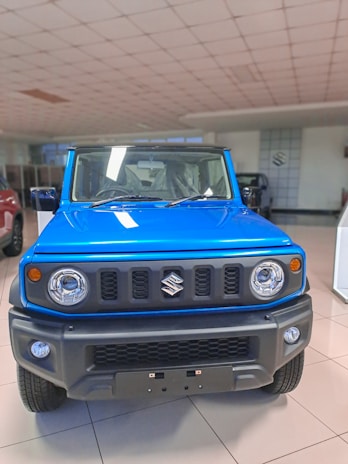 A vibrant blue SUV with a rugged front design is displayed in a well-lit showroom. The vehicle features round headlights, a bold grille with a logo, and an angular bumper. The floor is tiled, and other vehicles can be partially seen in the background.