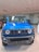 A vibrant blue SUV with a rugged front design is displayed in a well-lit showroom. The vehicle features round headlights, a bold grille with a logo, and an angular bumper. The floor is tiled, and other vehicles can be partially seen in the background.