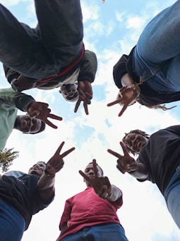 A group of people standing in a circle, looking down towards the camera while making peace signs with their hands. The sky is visible in the background, indicating the photo was taken from below.