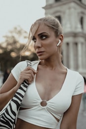 A person is wearing a white top with a ring detail in the front, accessorized with a zebra-patterned bag. They have wireless earbuds in their ears. The background suggests an urban setting with an architectural structure and trees.