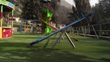 A playground featuring various play structures including a seesaw, a slide, and a climbing frame. The ground is covered in grass, and there are colorful railings and a canopy visible. Trees and a mountain are in the background, adding to the outdoor setting.