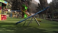 A playground featuring various play structures including a seesaw, a slide, and a climbing frame. The ground is covered in grass, and there are colorful railings and a canopy visible. Trees and a mountain are in the background, adding to the outdoor setting.