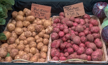 A display of organic potatoes with two varieties: Yukon Gold and Classic Red, each priced at $3.00 per pound. The Yukon Gold potatoes are tan and speckled, while the Classic Red potatoes are pinkish-red. Both types are placed in woven baskets. Nearby, there are leafy greens and cabbages.