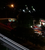 A nighttime urban scene featuring a roadway with light trails from moving vehicles, creating dynamic lines of red and white. A green highway sign indicates directions to Reagan National Airport. Streetlights illuminate parts of the scene with a warm glow, and a CVS pharmacy is visible on the right side with its bright signage.