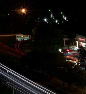 A nighttime urban scene featuring a roadway with light trails from moving vehicles, creating dynamic lines of red and white. A green highway sign indicates directions to Reagan National Airport. Streetlights illuminate parts of the scene with a warm glow, and a CVS pharmacy is visible on the right side with its bright signage.