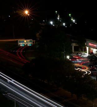 A nighttime urban scene featuring a roadway with light trails from moving vehicles, creating dynamic lines of red and white. A green highway sign indicates directions to Reagan National Airport. Streetlights illuminate parts of the scene with a warm glow, and a CVS pharmacy is visible on the right side with its bright signage.