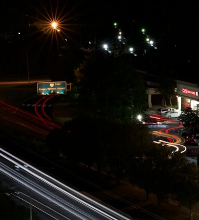 A nighttime urban scene featuring a roadway with light trails from moving vehicles, creating dynamic lines of red and white. A green highway sign indicates directions to Reagan National Airport. Streetlights illuminate parts of the scene with a warm glow, and a CVS pharmacy is visible on the right side with its bright signage.