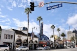 A street scene featuring a variety of buildings, including a prominent establishment marked with a large 'Hollywood' sign. Palm trees line the street, and there are several cars and people visible. The sky is partly cloudy, creating a sunny atmosphere. A street sign for McCadden Place is seen.