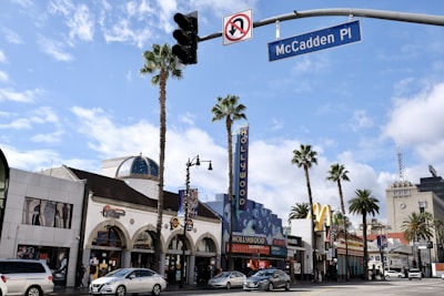 A street scene featuring a variety of buildings, including a prominent establishment marked with a large 'Hollywood' sign. Palm trees line the street, and there are several cars and people visible. The sky is partly cloudy, creating a sunny atmosphere. A street sign for McCadden Place is seen.