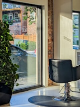 A modern interior with a salon chair facing large windows, allowing natural light to stream in. Outside, a car is parked near a brick building with green foliage and wooden accents visible through the glass. A large potted plant sits indoors by the window.
