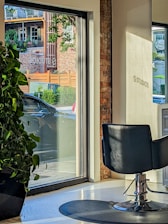A modern interior with a salon chair facing large windows, allowing natural light to stream in. Outside, a car is parked near a brick building with green foliage and wooden accents visible through the glass. A large potted plant sits indoors by the window.