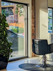 A modern interior with a salon chair facing large windows, allowing natural light to stream in. Outside, a car is parked near a brick building with green foliage and wooden accents visible through the glass. A large potted plant sits indoors by the window.