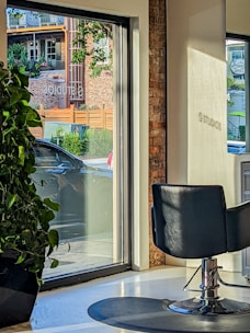 A modern interior with a salon chair facing large windows, allowing natural light to stream in. Outside, a car is parked near a brick building with green foliage and wooden accents visible through the glass. A large potted plant sits indoors by the window.