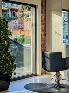 A modern interior with a salon chair facing large windows, allowing natural light to stream in. Outside, a car is parked near a brick building with green foliage and wooden accents visible through the glass. A large potted plant sits indoors by the window.