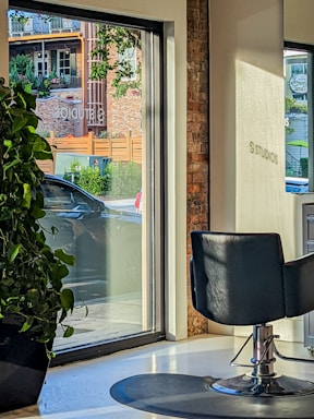 A modern interior with a salon chair facing large windows, allowing natural light to stream in. Outside, a car is parked near a brick building with green foliage and wooden accents visible through the glass. A large potted plant sits indoors by the window.