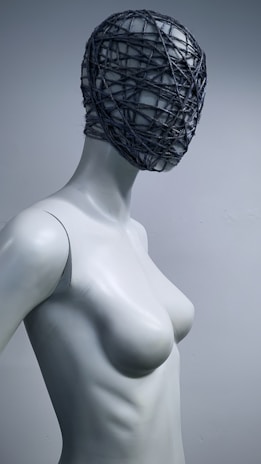A mannequin with a smooth, featureless surface and a head wrapped in dark, string-like material. The setting shows a simplistic and minimalist style with a monochromatic color scheme.