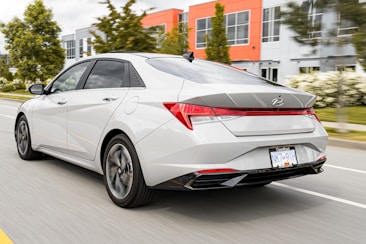 A modern sedan is driving on a road with blurred green trees and contemporary architectural buildings in the background, indicating movement and speed. The car is white with sleek, aerodynamic features, emphasized by its angular design and rear spoiler.