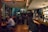 A dimly lit bar with groups of people sitting at tables and the bar counter, engaging in conversation and enjoying drinks. The room has a cozy ambiance with warm lighting and an eclectic mix of posters on the walls. The bar area is crowded, and the patrons appear relaxed and social.