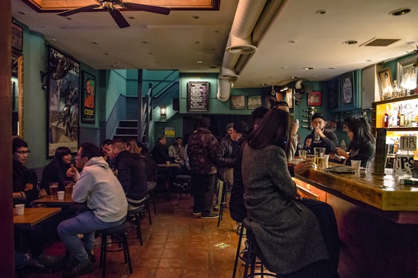 A dimly lit bar with groups of people sitting at tables and the bar counter, engaging in conversation and enjoying drinks. The room has a cozy ambiance with warm lighting and an eclectic mix of posters on the walls. The bar area is crowded, and the patrons appear relaxed and social.