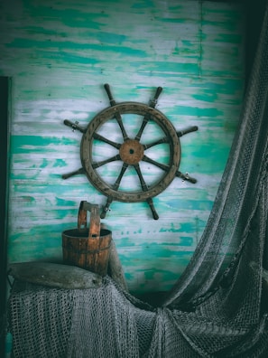 A rustic ship's wheel is mounted on a weathered, teal and white wooden wall. Below it, a wooden barrel is surrounded by nautical ropes and a fishing net.