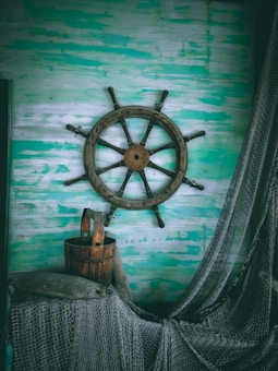 A rustic ship's wheel is mounted on a weathered, teal and white wooden wall. Below it, a wooden barrel is surrounded by nautical ropes and a fishing net.