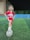 A young girl is standing on a soccer field at night. She is wearing a red soccer jersey, white sneakers, and has her foot on a soccer ball. The field is lined with bright green artificial grass, surrounded by blue barriers and a fence. A building is visible in the background.