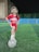 A young girl is standing on a soccer field at night. She is wearing a red soccer jersey, white sneakers, and has her foot on a soccer ball. The field is lined with bright green artificial grass, surrounded by blue barriers and a fence. A building is visible in the background.