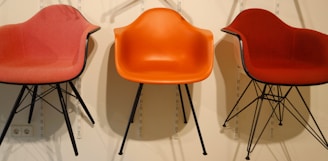 Three modern chairs with distinct seating colors, mounted against a light yellow wall. The chairs have a smooth, curved design with black metal legs. The central chair is a bright orange, while the two on the sides are shades of red.