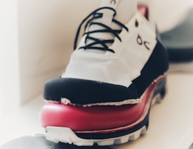 A close-up view of a sneaker with a distinct design featuring a layered sole. The shoe has a combination of white, black, and red colors, with black laces and a visible logo on the side. The focus is on the side of the sneaker, highlighting the texture and unique layering of its sole. The background is blurred, drawing attention to the shoe.