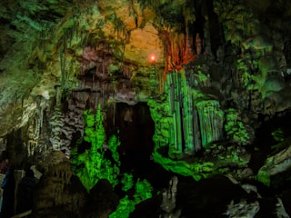 A breathtaking view of a cave interior featuring striking stalactites and stalagmites illuminated with green and orange lighting, creating a dramatic and mystical atmosphere. The texture of the rock formations is intricate and rugged, with shadows accentuating their form.