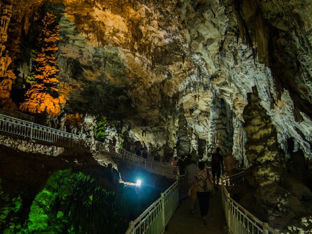 An illuminated underground cave with stalactites and stalagmites showcasing intricate rock formations. A pathway with railings allows visitors to explore the cave. The lighting casts dramatic shadows and highlights the textured surfaces of the rocks.