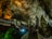 An illuminated underground cave with stalactites and stalagmites showcasing intricate rock formations. A pathway with railings allows visitors to explore the cave. The lighting casts dramatic shadows and highlights the textured surfaces of the rocks.