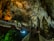 An illuminated underground cave with stalactites and stalagmites showcasing intricate rock formations. A pathway with railings allows visitors to explore the cave. The lighting casts dramatic shadows and highlights the textured surfaces of the rocks.