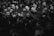 A large group of people gathered closely together, with a range of emotional expressions visible on their faces. Some individuals are visibly upset or crying, while others look pensive or sad. A few people are wearing masks, and most are dressed in dark clothing. The setting appears to be a crowded indoor space with dim lighting.
