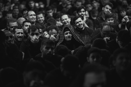 A large group of people gathered closely together, with a range of emotional expressions visible on their faces. Some individuals are visibly upset or crying, while others look pensive or sad. A few people are wearing masks, and most are dressed in dark clothing. The setting appears to be a crowded indoor space with dim lighting.