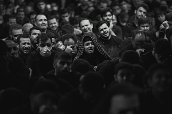 A large group of people gathered closely together, with a range of emotional expressions visible on their faces. Some individuals are visibly upset or crying, while others look pensive or sad. A few people are wearing masks, and most are dressed in dark clothing. The setting appears to be a crowded indoor space with dim lighting.
