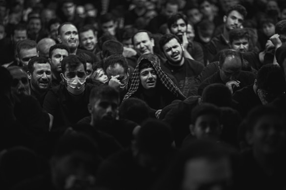 A large group of people gathered closely together, with a range of emotional expressions visible on their faces. Some individuals are visibly upset or crying, while others look pensive or sad. A few people are wearing masks, and most are dressed in dark clothing. The setting appears to be a crowded indoor space with dim lighting.