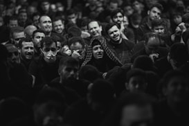 A large group of people gathered closely together, with a range of emotional expressions visible on their faces. Some individuals are visibly upset or crying, while others look pensive or sad. A few people are wearing masks, and most are dressed in dark clothing. The setting appears to be a crowded indoor space with dim lighting.