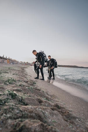 Two scuba divers in full gear walk on a sandy beach adjacent to the sea. The shoreline is visible with calm waters and a clear sky overhead. There are beach structures and umbrellas in the distance.