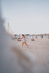 A person is playing beach volleyball on a sandy beach. Other people are lounging and walking in the background. The sky is clear and the beach umbrellas are visible in the distance.
