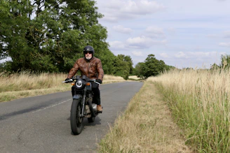 A biker wearing a brown leather jacket and gloves, gripping the handlebar on an open road.