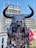 A massive mechanical sculpture of a bull with intricate metallic features and large horns stands prominently with a modern building in the background. The bull has a nose ring and chains hanging from its mouth, and its design is industrial and artistic.