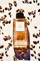 A bottle of D&G 11 La Force perfume is positioned in the center, surrounded by scattered cardamom pods on a white surface. The bottle is filled with an amber-colored liquid and features a black cap.
