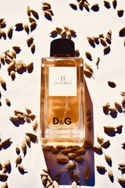 A bottle of D&G 11 La Force perfume is positioned in the center, surrounded by scattered cardamom pods on a white surface. The bottle is filled with an amber-colored liquid and features a black cap.