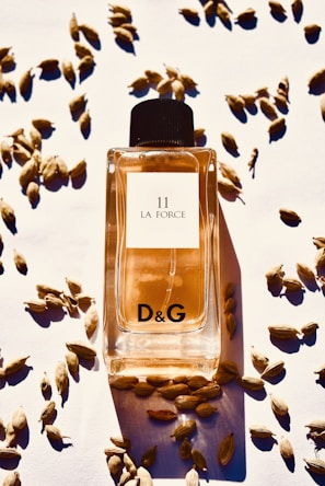 A bottle of D&G 11 La Force perfume is positioned in the center, surrounded by scattered cardamom pods on a white surface. The bottle is filled with an amber-colored liquid and features a black cap.