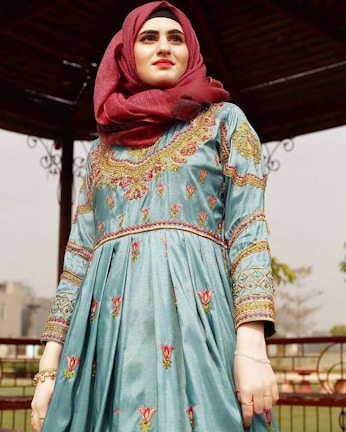A person is wearing a traditional embroidered dress featuring intricate floral patterns and geometric designs. The dress is complemented by a maroon headscarf. The background includes elements of a wooden gazebo and some greenery, creating a serene outdoor setting.