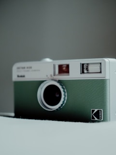 A vintage-style camera with a green leather texture on a neutral background. The camera has a distinctive circular lens and a small viewfinder, with branding on the front panel.